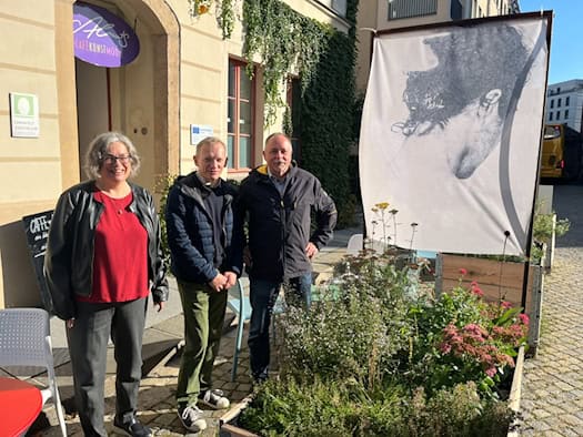 Drei Personen – Eva Jähnigen, Tom Umbreit und André Barth – stehen lächelnd vor einem bepflanzten Holzhochbeet und einem Kunstbanner neben dem Eingang des Umweltzentrums Dresden.