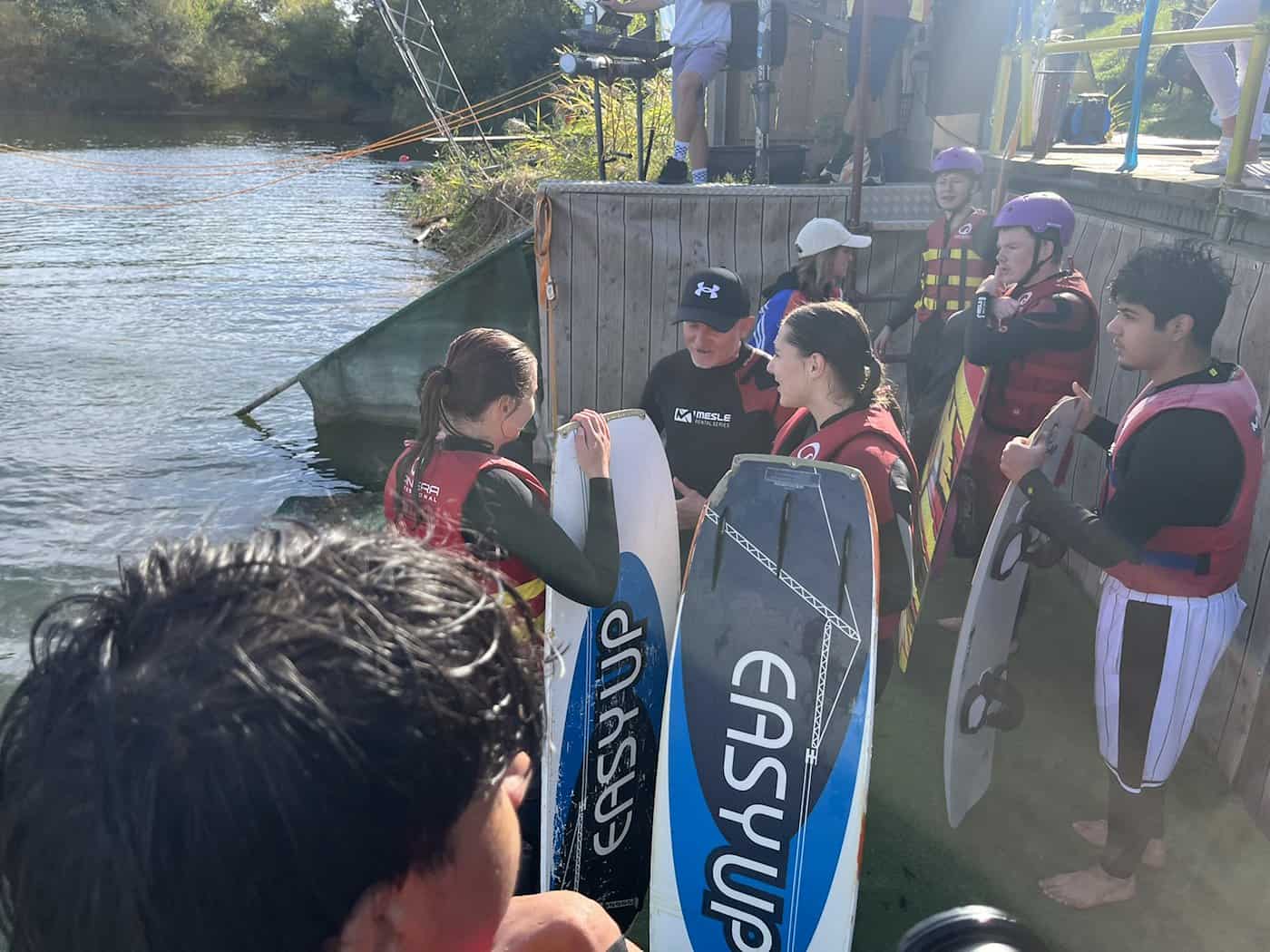 Jugendliche in Neoprenanzügen und Schwimmwesten stehen mit Wakeboards am Holzsteg und hören aufmerksam ihrem Betreuer zu. Im Vordergrund sind Wakeboards zu sehen.