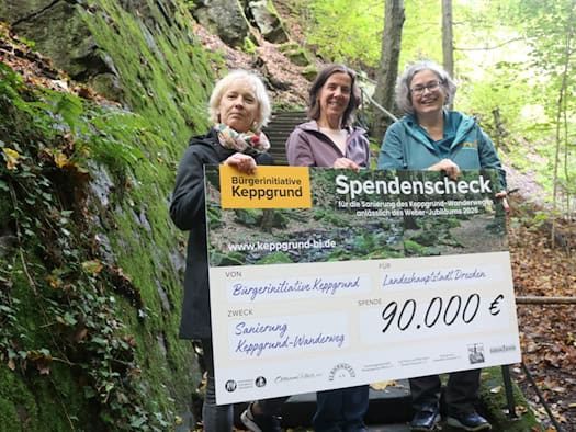 Drei Frauen – Simone Prüfer, Manuela Schreiter und Eva Jähnigen (v.l.) – stehen lächelnd auf einer Steintreppe im Keppgrund und halten gemeinsam einen großen Spendenscheck der Bürgerinitiative über 90.000 Euro in die Kamera.