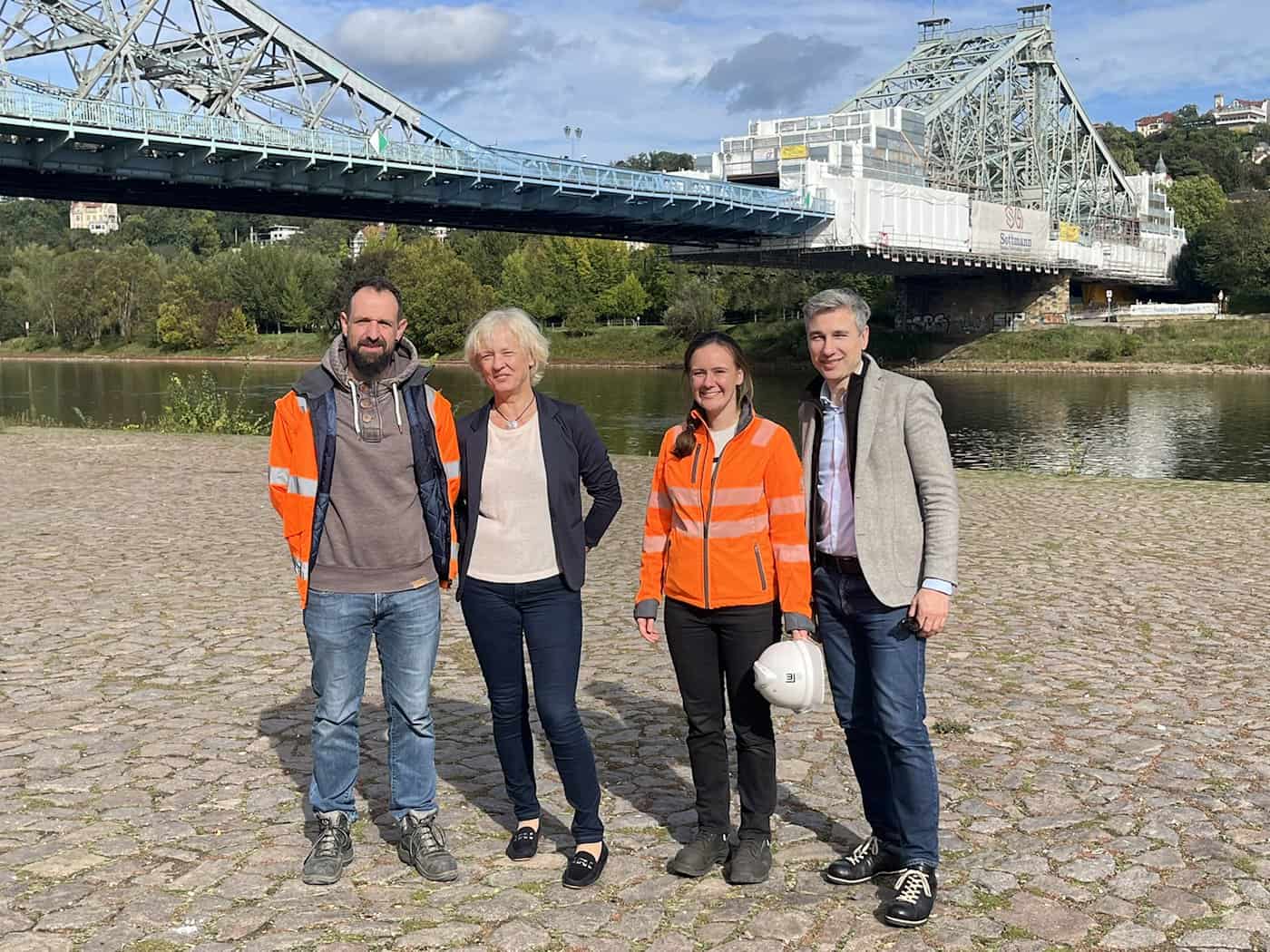Vier Personen stehen nebeneinander am Elbufer vor der stählernen Fachwerksbrücke Blaues Wunder in Dresden, die teilweise eingerüstet ist. Im Hintergrund fließt die Elbe unter blauem Himmel.