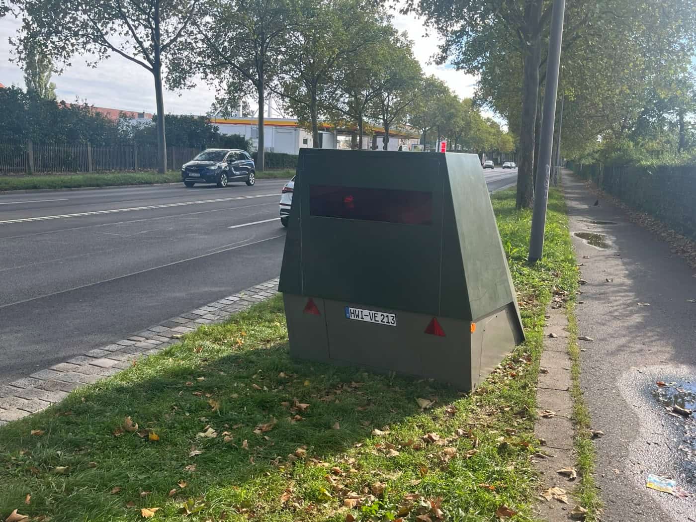 Ein olivgrüner, panzerartiger Messanhänger mit dem Nummernschild HWI-VE 213 steht auf einem Grünstreifen an einer mehrspurigen Straße neben einem Fußweg. Im Hintergrund sind fahrende Autos und eine Tankstelle zu sehen.