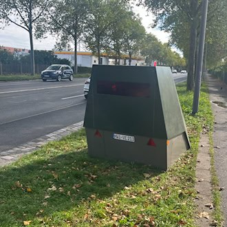 Ein olivgrüner, panzerartiger Messanhänger mit dem Nummernschild HWI-VE 213 steht auf einem Grünstreifen an einer mehrspurigen Straße neben einem Fußweg. Im Hintergrund sind fahrende Autos und eine Tankstelle zu sehen.
