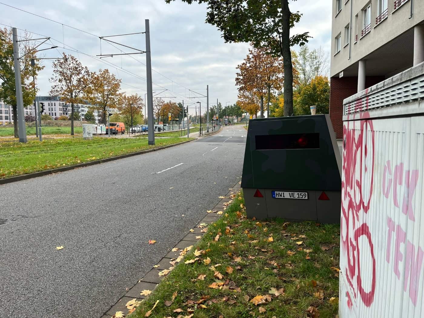 Nahaufnahme des grauen Mess-Anhängers mit dem blauen EU-Kfz-Kennzeichen „HWI-VE 159“ und rot-weißen Warndreiecken; davor steht ein mit Graffiti beschmierter grauer Verteilerkasten im herbstlichen Laub.