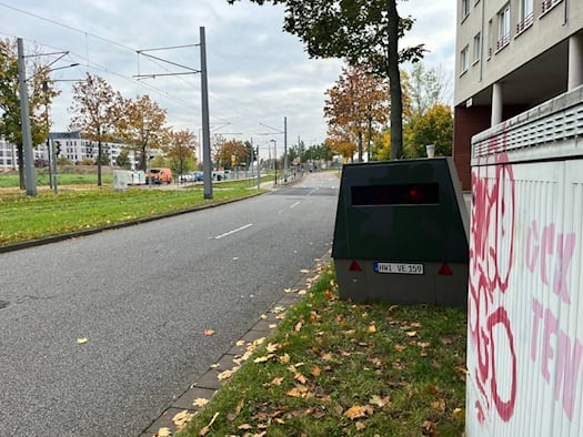 Nahaufnahme des grauen Mess-Anhängers mit dem blauen EU-Kfz-Kennzeichen „HWI-VE 159“ und rot-weißen Warndreiecken; davor steht ein mit Graffiti beschmierter grauer Verteilerkasten im herbstlichen Laub.