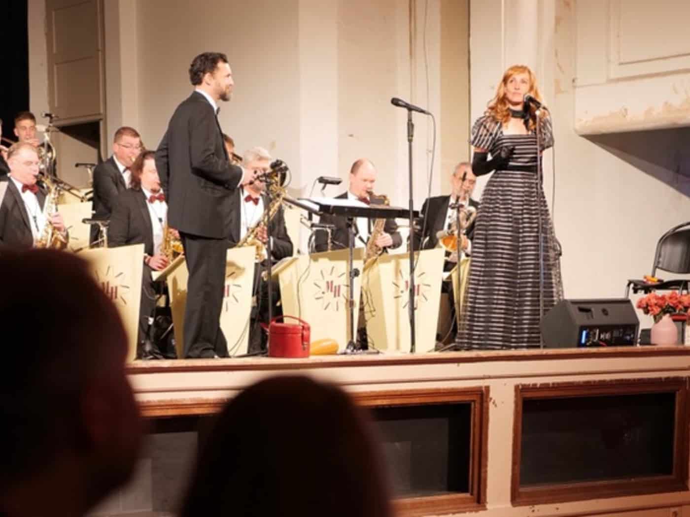 Sängerin im gestreiften Kleid und Bandleader im Frack auf der Bühne im Henny-Brenner-Saal des Zentralwerks. Dahinter spielt Marc Hartmanns Tanzorchester in Abendgarderobe mit Blasinstrumenten. Blick aus dem Publikum auf die erhöhte Bühne.