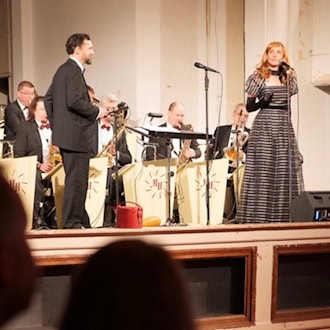 Sängerin im gestreiften Kleid und Bandleader im Frack auf der Bühne im Henny-Brenner-Saal des Zentralwerks. Dahinter spielt Marc Hartmanns Tanzorchester in Abendgarderobe mit Blasinstrumenten. Blick aus dem Publikum auf die erhöhte Bühne.