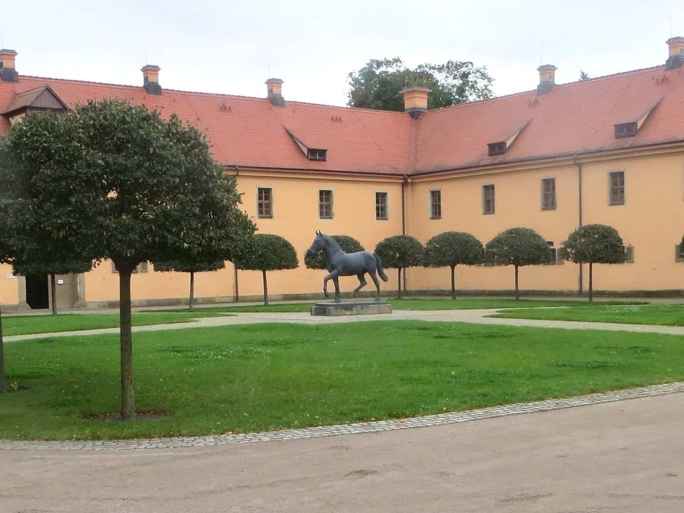 Innenhof des Landgestüts Moritzburg mit Rasen, Kugelbäumen und Pferdestatue vor gelben Stallgebäuden