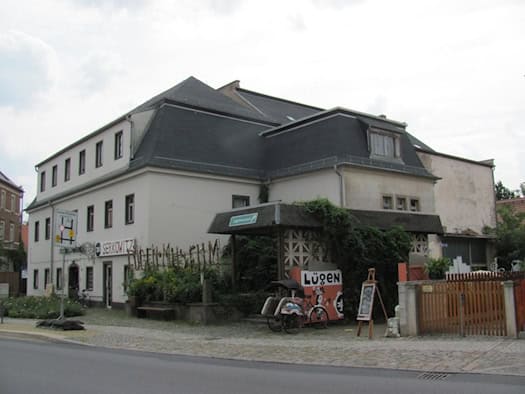Zweistöckiges weißes Gasthaus mit dunklem Mansarddach an einer Straße, davor ein Vordach mit Efeu, ein Schild „Lügen“ und ein Rikscha-Fahrrad