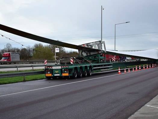 Ein langes Rotorblatt einer Windkraftanlage liegt auf einem Schwertransporter auf einem Autobahnparkplatz.