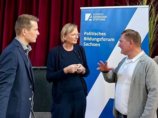 Daniela Kuge im Gespräch mit zwei Männern vor einem Aufsteller des Politischen Bildungsforums Sachsen der Konrad-Adenauer-Stiftung