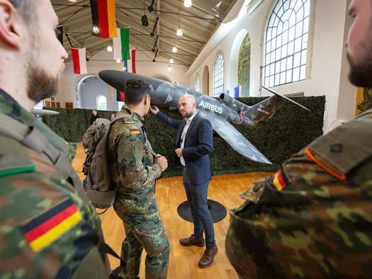 Soldaten in Flecktarnuniform stehen in einer Halle mit Flaggen vor einem großen Drohnenmodell mit Airbus-Schriftzug, ein Mann im Anzug erklärt es.