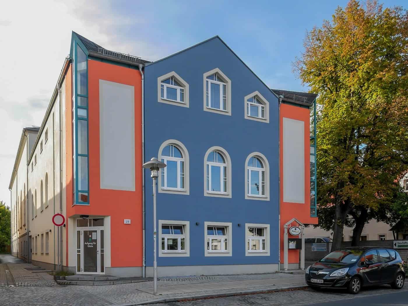 Die Börse Coswig an der Hauptstraße 29 mit blauer Fassade und orangefarbenen Seitenteilen, rechts ein Laubbaum in Herbstfarben.