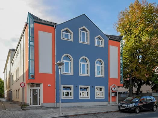Die Börse Coswig an der Hauptstraße 29 mit blauer Fassade und orangefarbenen Seitenteilen, rechts ein Laubbaum in Herbstfarben.