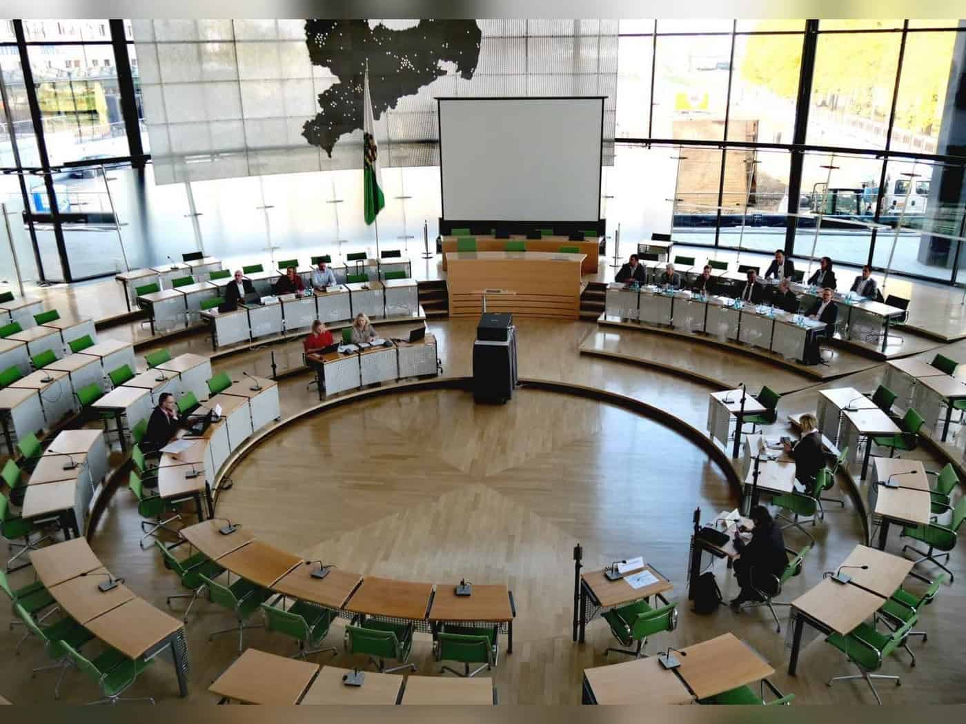 Blick von oben in den Plenarsaal des Sächsischen Landtags mit Holztischen, grünen Stühlen und mehreren Personen an den Tischen