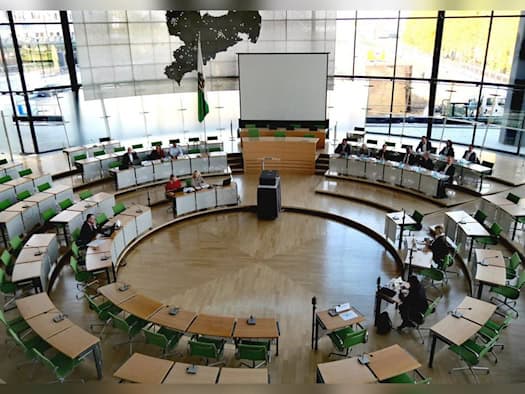 Blick von oben in den Plenarsaal des Sächsischen Landtags mit Holztischen, grünen Stühlen und mehreren Personen an den Tischen