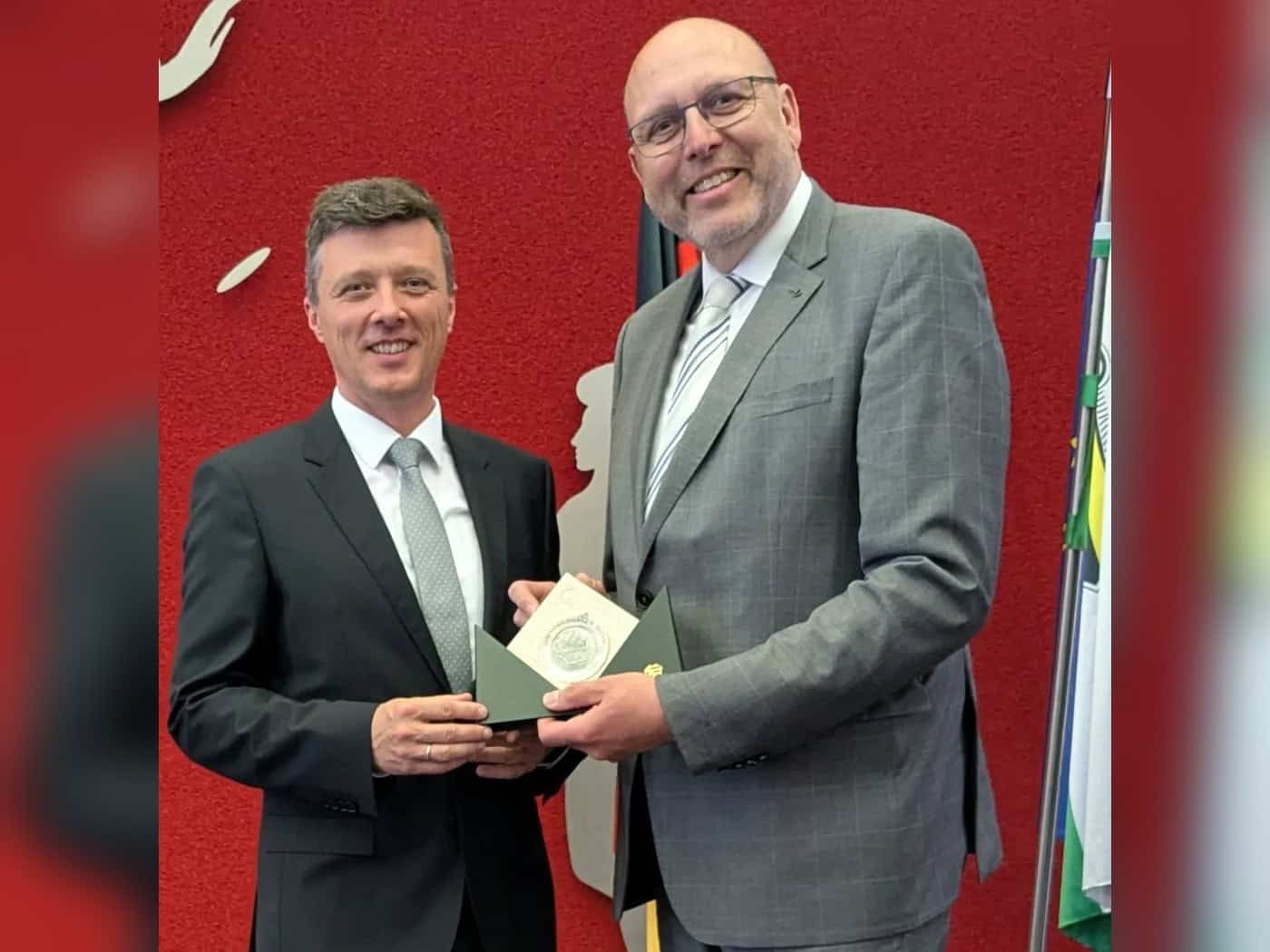Markus Renner (links) und Olaf Raschke lächeln in die Kamera und halten gemeinsam eine aufgeklappte grüne Mappe mit einer Plakette.