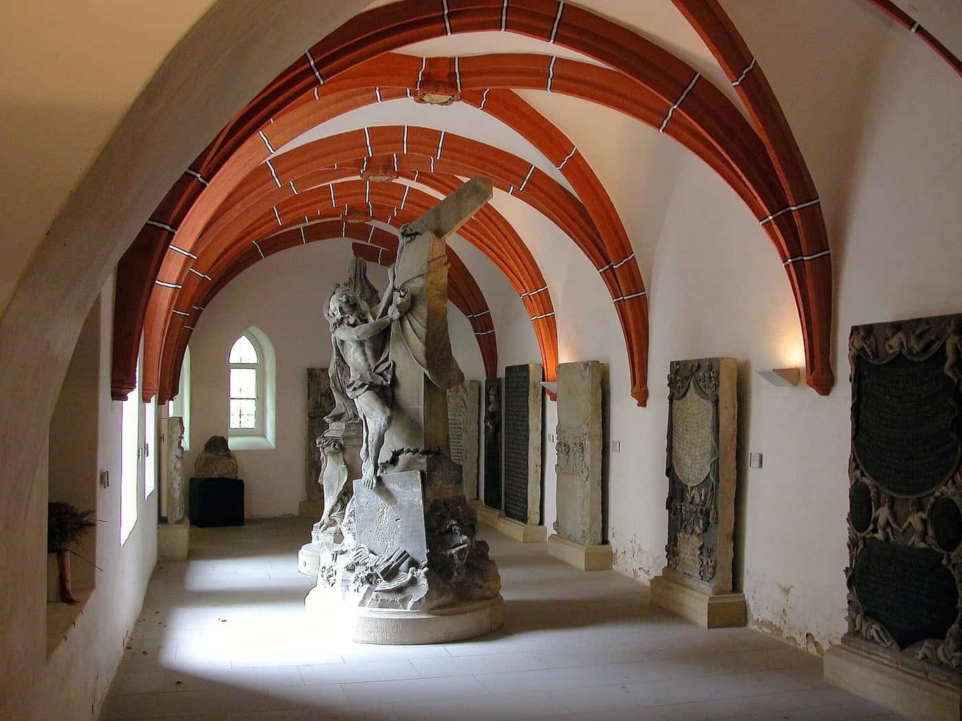 Gewölbter Ausstellungsgang mit roten Gewölberippen, darin eine Steinskulptur und historische Grabplatten an den Wänden