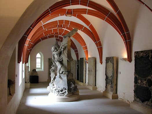 Gewölbter Ausstellungsgang mit roten Gewölberippen, darin eine Steinskulptur und historische Grabplatten an den Wänden
