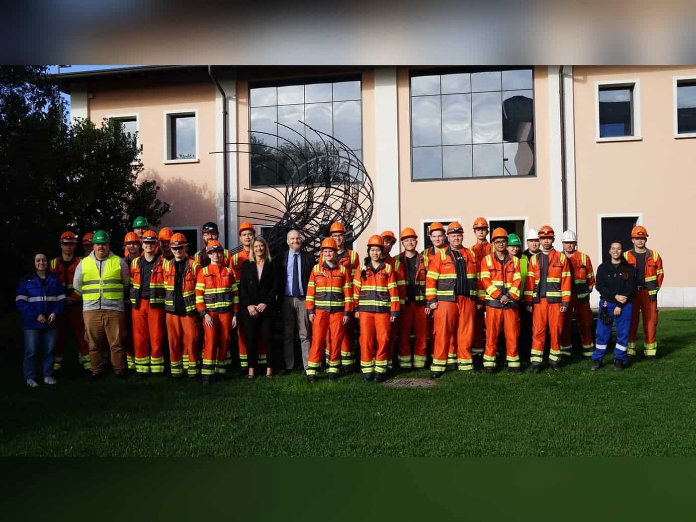 Gruppe von Auszubildenden und Studenten in orangefarbener Arbeitskleidung mit Schutzhelmen vor einem Gebäude in Italien