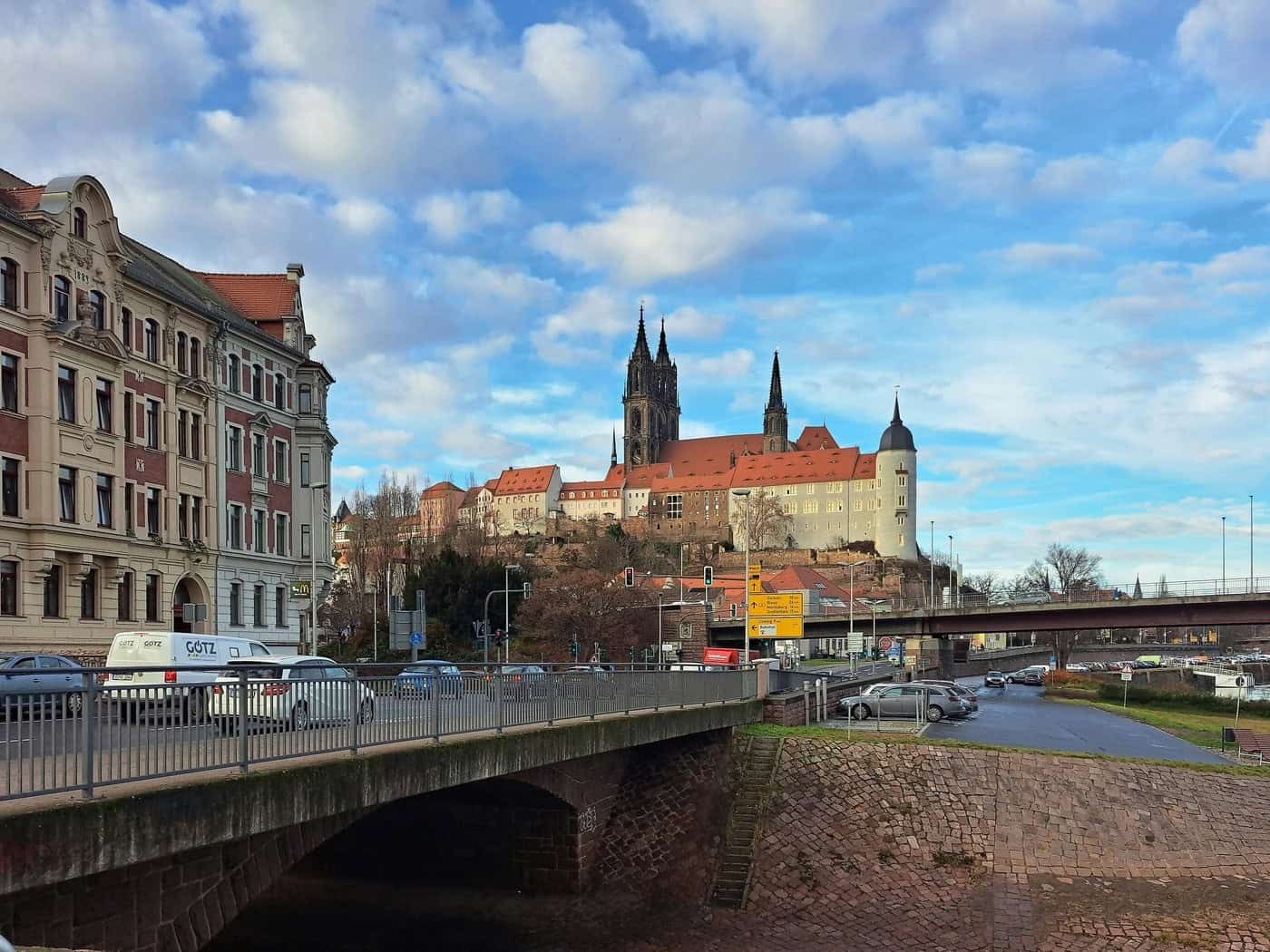 Blick von einer Brücke über Häuser an der Elbe auf die Meißner Albrechtsburg und die beiden Domtürme unter bewölktem Himmel.