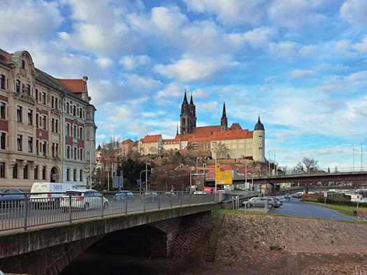 Blick von einer Brücke über Häuser an der Elbe auf die Meißner Albrechtsburg und die beiden Domtürme unter bewölktem Himmel.