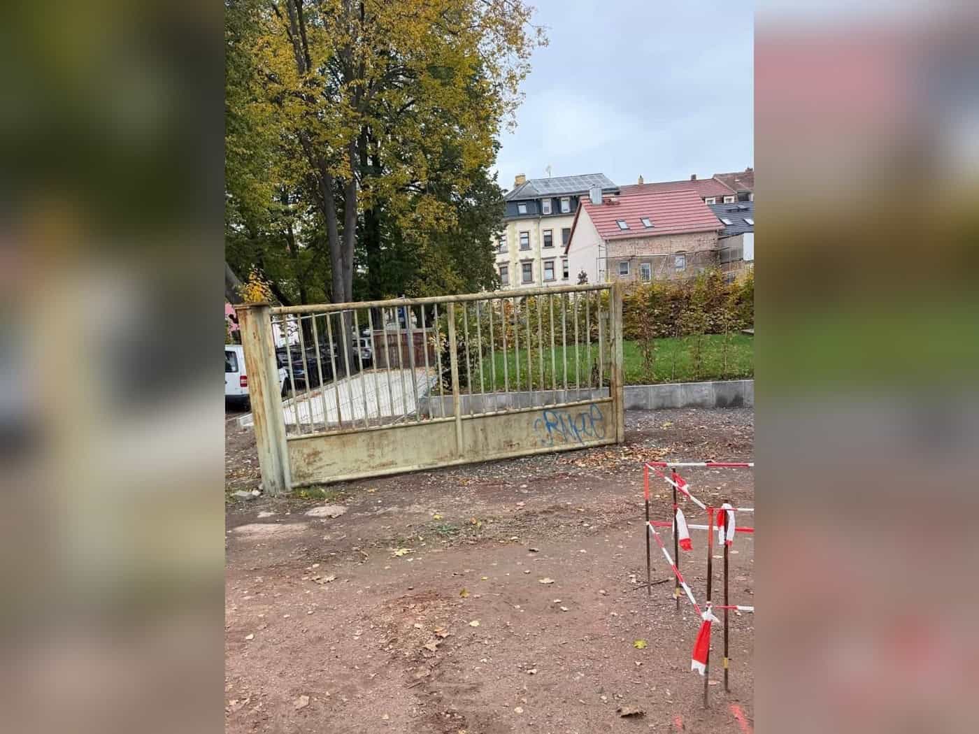 Rostiges, hellgrünes Metalltor mit senkrechten Streben liegt auf einem Erdplatz, dahinter Herbstbäume und Wohnhäuser.