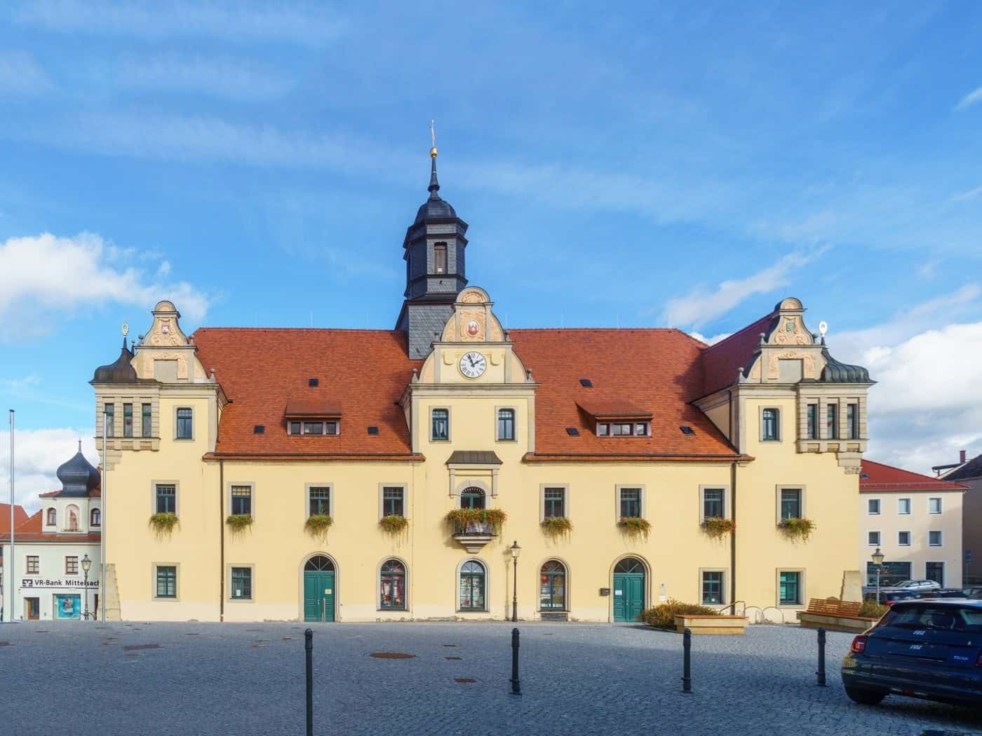 Das gelbe Rathaus von Lommatzsch mit rotem Dach und Uhrenturm, davor der gepflasterte Marktplatz