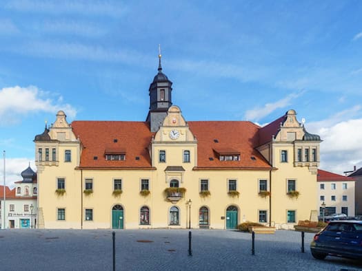 Das gelbe Rathaus von Lommatzsch mit rotem Dach und Uhrenturm, davor der gepflasterte Marktplatz