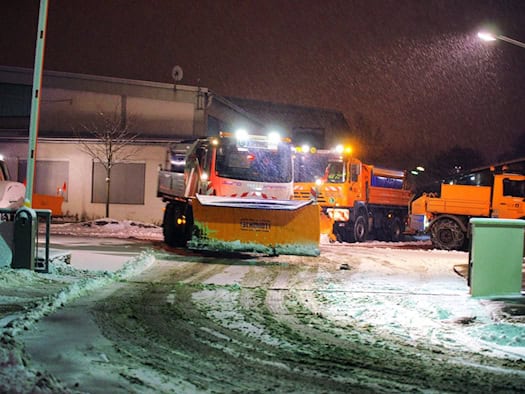 Orangefarbene Räumfahrzeuge mit Schneeschild fahren nachts bei Schneefall aus einem Betriebshof
