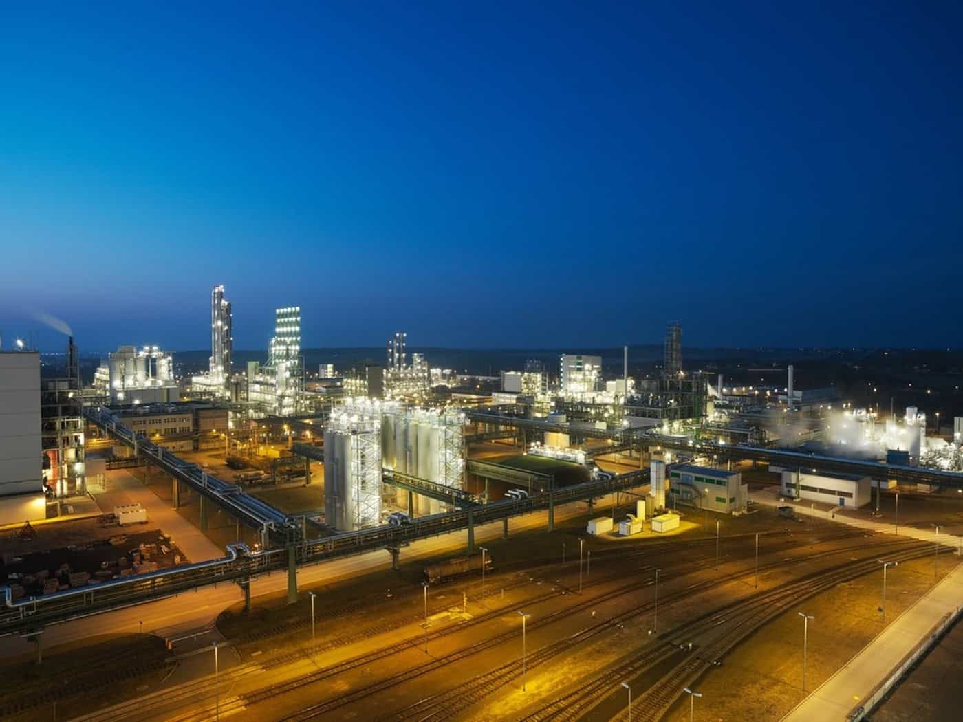 Beleuchtetes Chemiewerk mit Produktionsanlagen, Rohrbrücken und Gleisen in der Dämmerung