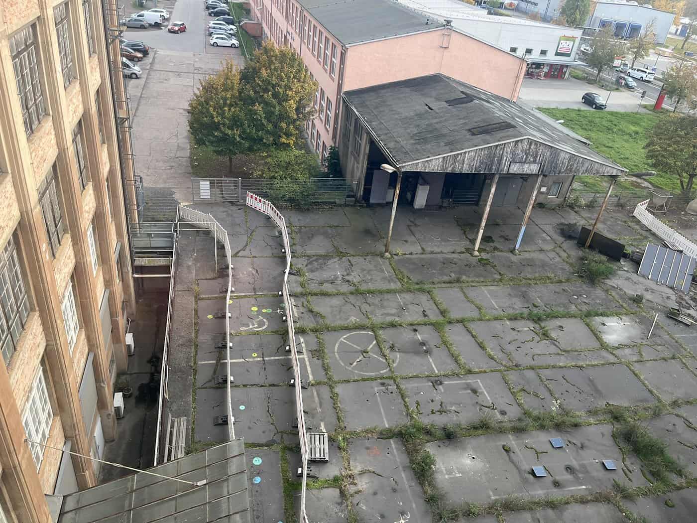Draufsicht aus großer Höhe auf den asphaltierten Innenhof der Technischen Sammlungen Dresden mit Markierungen, Bauzäunen und Schuppenanbauten. Links ragt die hohe Backsteinfassade des Hauptgebäudes in das Bild.