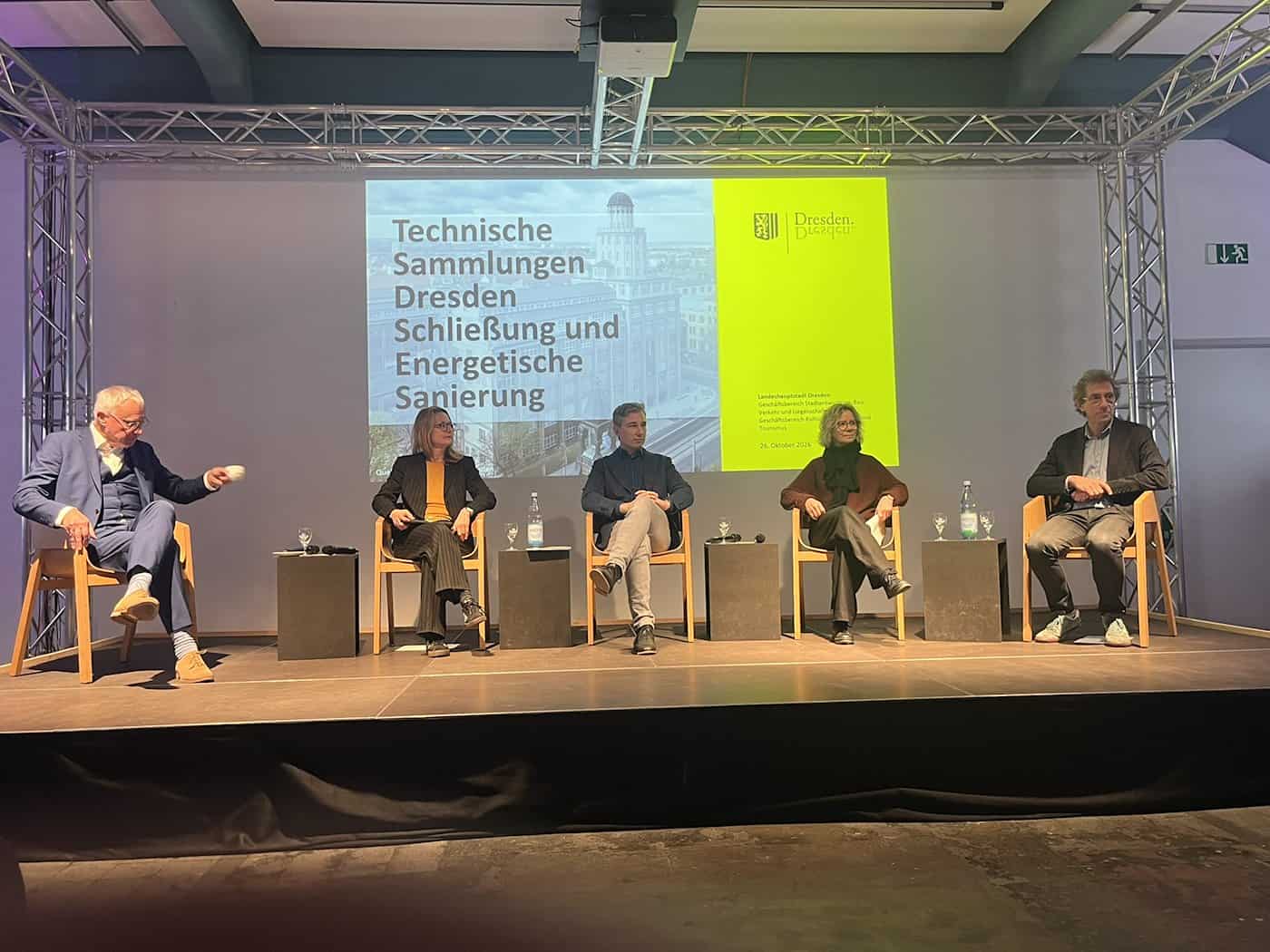 Fünf Personen sitzen bei einer Pressekonferenz auf Stühlen auf einer Bühne. Im Hintergrund zeigt eine Projektionsleinwand die Präsentation „Technische Sammlungen Dresden Schließung und Energetische Sanierung“ mit dem Logo der Landeshauptstadt Dresden.