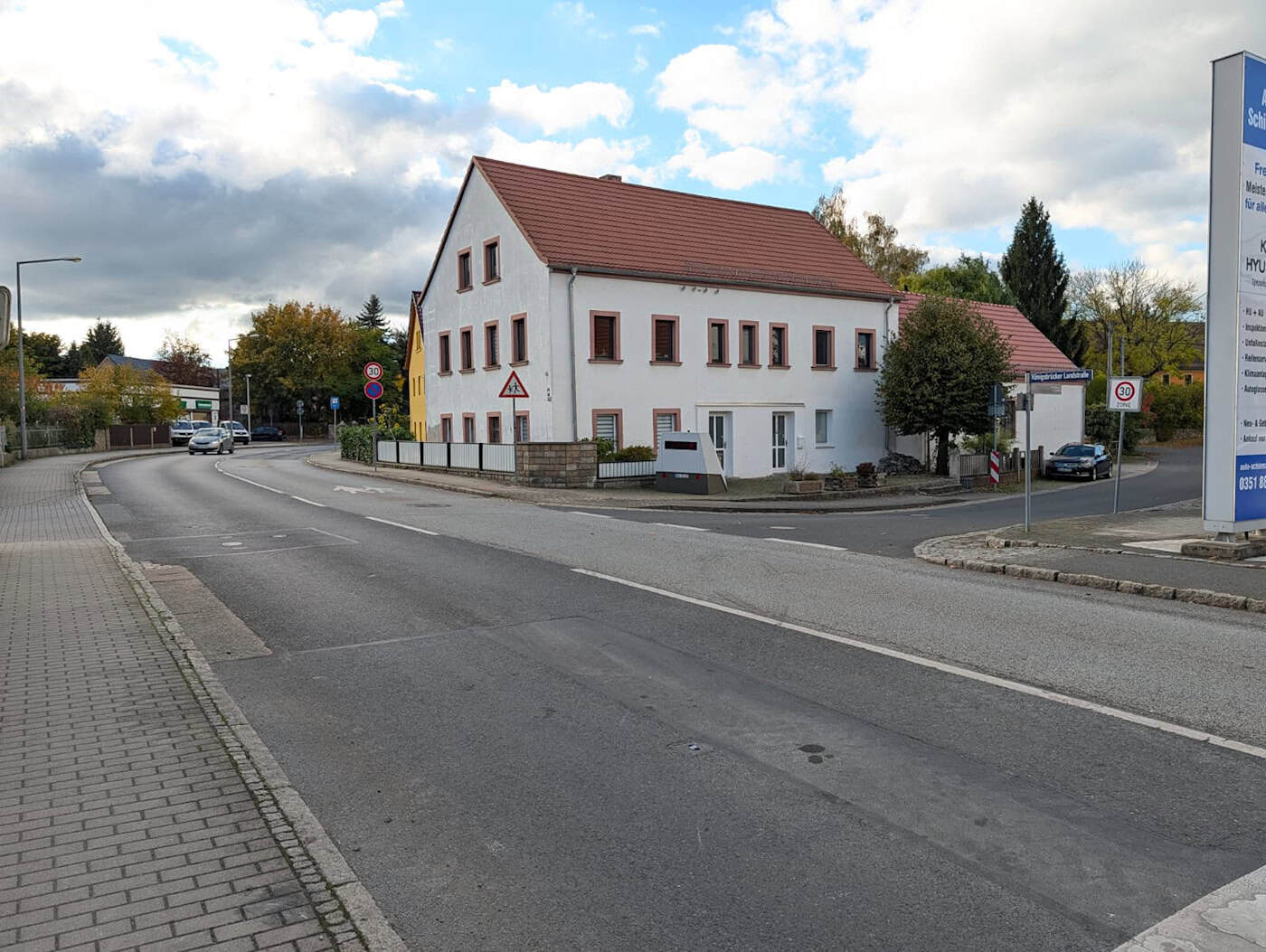 Totalansicht der Kreuzung in Weixdorf mit dem aufgestellten Superblitzer Weitwinkelblick auf die Straße und Kreuzung in Dresden-Weixdorf unter teils bewölktem Himmel; der graue Blitzer-Anhänger steht an einem weißen Wohnhaus, während Autos auf der Straße unterwegs sind.