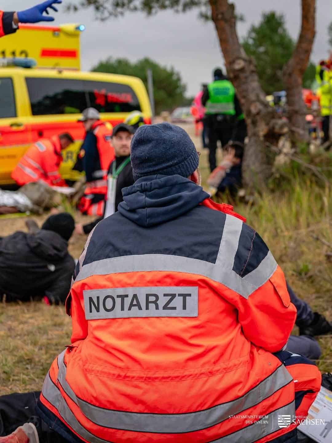 Ein Notarzt in orangener Jacke sitzt bei Verletzten auf einer Wiese, im Hintergrund ein Rettungswagen