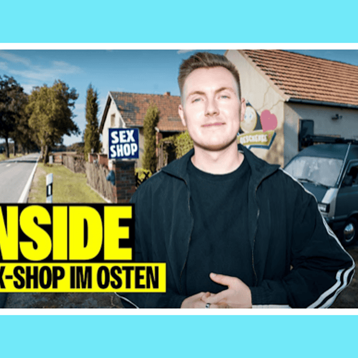 YouTuber Tomatolix steht lächelnd an einer Landstraße vor einem Gebäude mit blauem Schild „Sex Shop“, daneben parkt ein alter Bus; gelb-schwarzer Schriftzug-Einblender „INSIDE SEX-SHOP IM OSTEN“.