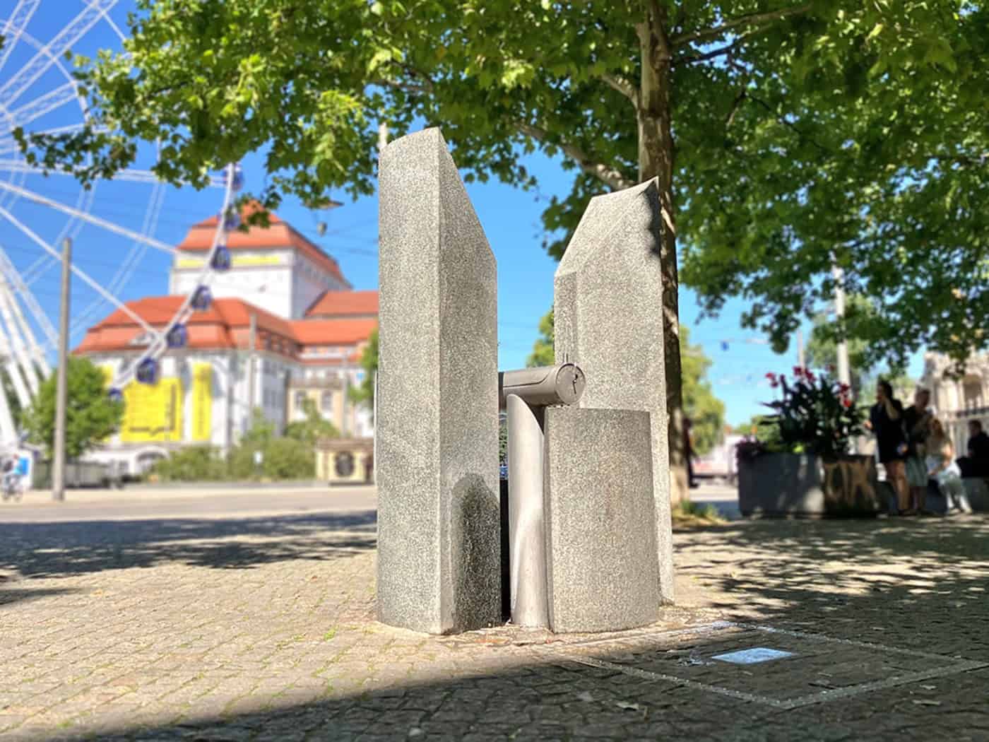 Ein moderner Trinkwasserbrunnen aus Stein und Edelstahl auf gepflastertem Platz bei Sonnenschein; im unscharfen Hintergrund sind ein Riesenrad, das Schauspielhaus Dresden und grüne Bäume zu sehen.