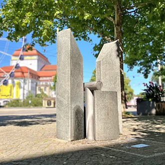 Ein moderner Trinkwasserbrunnen aus Stein und Edelstahl auf gepflastertem Platz bei Sonnenschein; im unscharfen Hintergrund sind ein Riesenrad, das Schauspielhaus Dresden und grüne Bäume zu sehen.