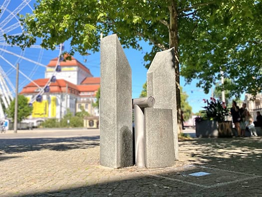 Ein moderner Trinkwasserbrunnen aus Stein und Edelstahl auf gepflastertem Platz bei Sonnenschein; im unscharfen Hintergrund sind ein Riesenrad, das Schauspielhaus Dresden und grüne Bäume zu sehen.