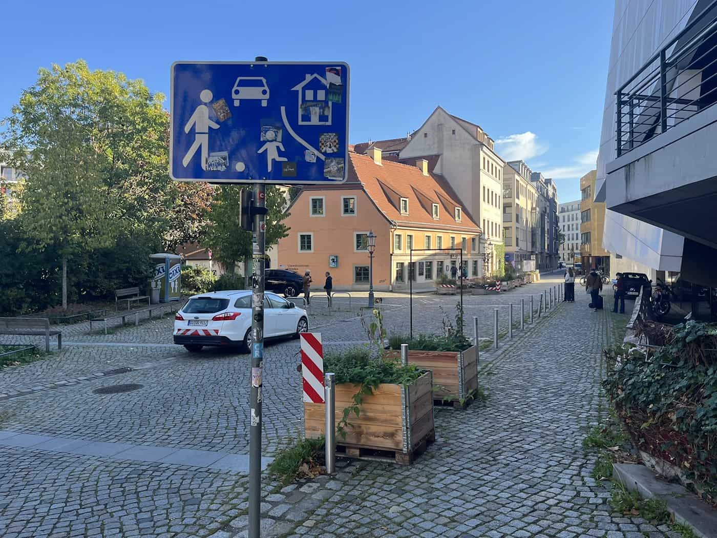 Nahaufnahme eines blauen Straßenschilds für einen verkehrsberuhigten Bereich; im Hintergrund ist die Schützengasse mit Kopfsteinpflaster, parkenden Autos und Holz-Pflanzkübeln zu sehen.
