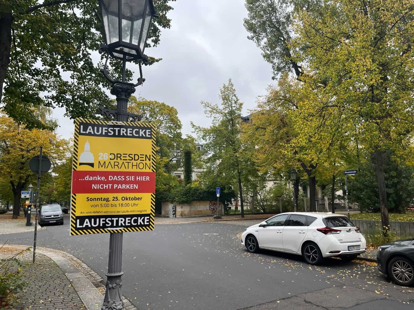 An einer historischen Straßenlaterne montiertes gelb-rotes Hinweisschild mit der Aufschrift „Laufstrecke 26. Dresden Marathon - danke, DASS SIE HIER NICHT PARKEN“ am Sonntag, 25. Oktober; im Hintergrund herbstliche Bäume und parkende Autos.