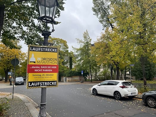 An einer historischen Straßenlaterne montiertes gelb-rotes Hinweisschild mit der Aufschrift „Laufstrecke 26. Dresden Marathon - danke, DASS SIE HIER NICHT PARKEN“ am Sonntag, 25. Oktober; im Hintergrund herbstliche Bäume und parkende Autos.
