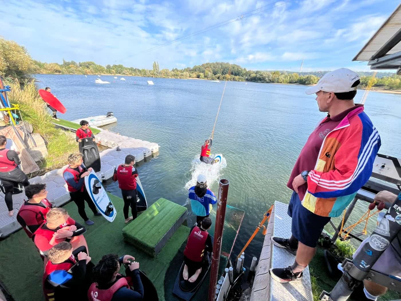 Blick von oben auf den Kiessee Leuben: Ein Schüler startet am Seilzug auf dem Wasser, während am Steg Mitschüler in Schwimmwesten und ein Betreuer in bunter Jacke das Geschehen beobachten.