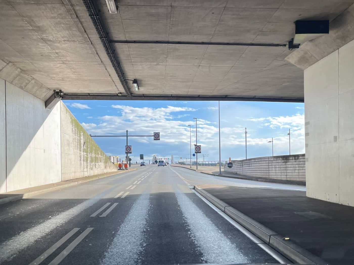 Blick aus der Windschutzscheibe eines Autos bei der Ausfahrt aus der hellgrauen Weströhre des Waldschlößchentunnels in Dresden auf die Fahrbahn in Richtung Waldschlösschenbrücke unter blauem, leicht bewölktem Himmel.