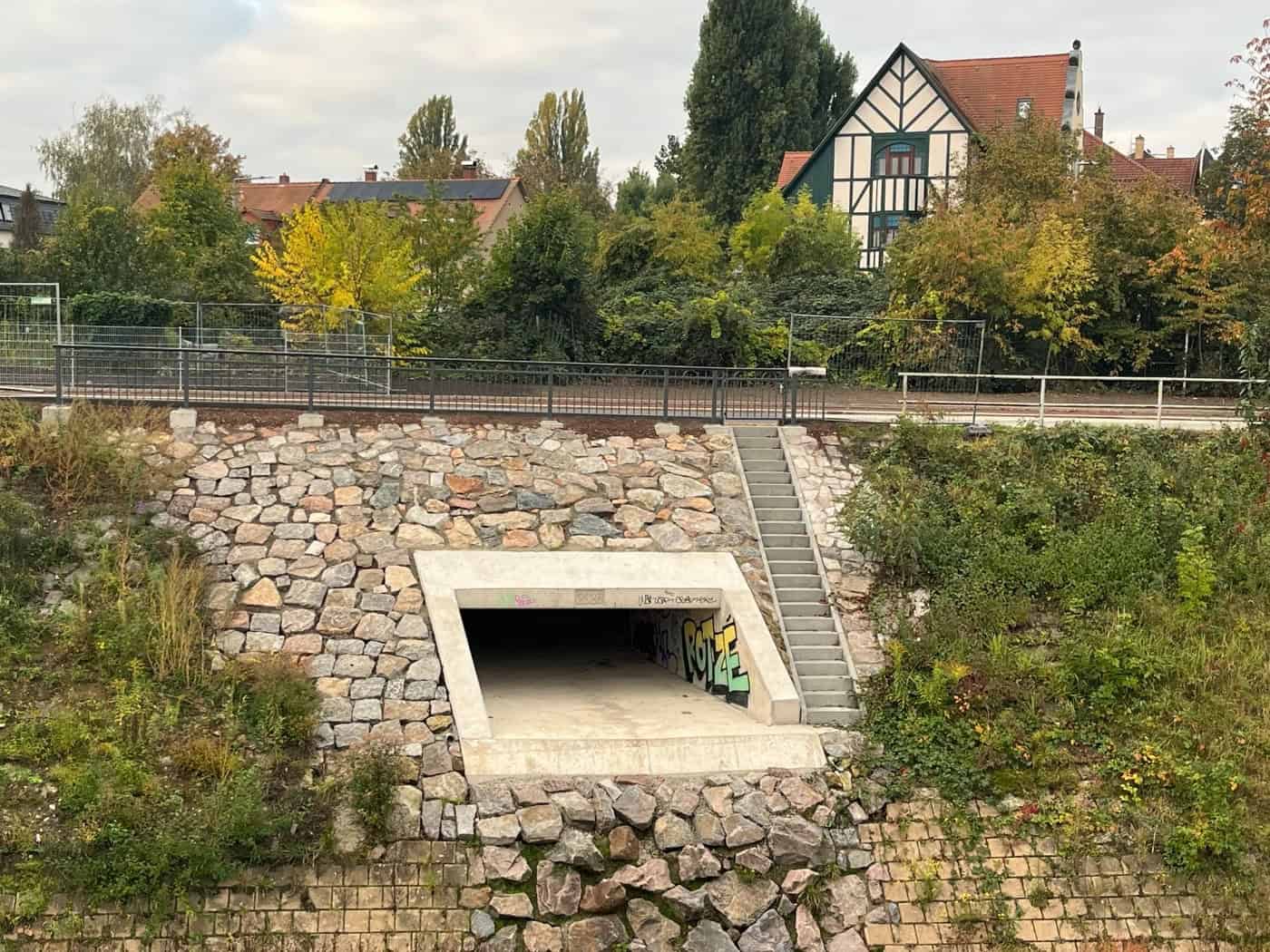 Neu errichteter Beton-Durchlass für den Weidigtbach mit Naturstein-Böschung und Betontreppe; darüber verläuft das Geländer des Emerich-Ambros-Ufers mit herbstlichen Bäumen und Fachwerkhäusern im Hintergrund.
