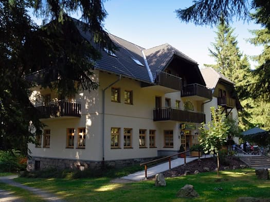 ein Gebäude mit 2 Etagen umgeben von Wald, am Eingang ein Restaurantschild, davor ein Freisitz mit Gästen