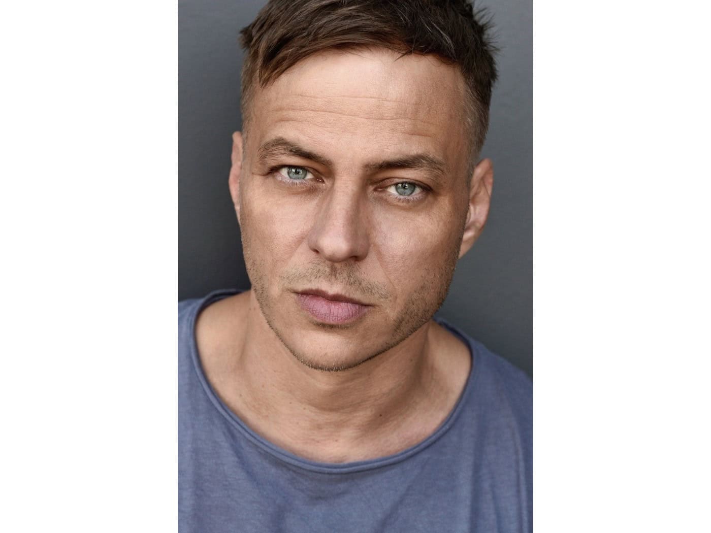 Porträt des Schauspielers Tom Wlaschiha mit kurzen Haaren und grau-blauem T-Shirt vor grauem Hintergrund