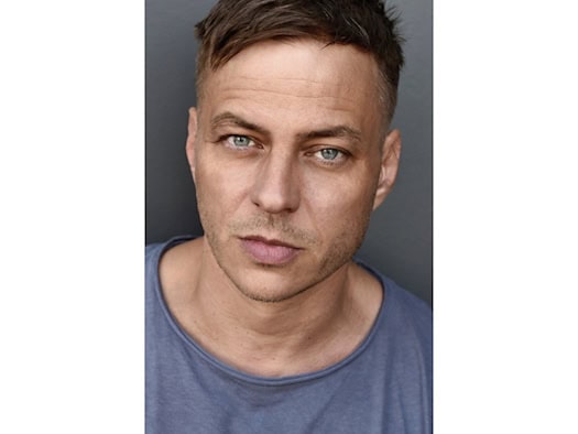 Porträt des Schauspielers Tom Wlaschiha mit kurzen Haaren und grau-blauem T-Shirt vor grauem Hintergrund
