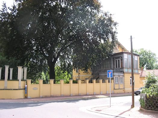 Gelbe Villa hinter einer Mauer mit Pfeilern, davor ein großer Laubbaum