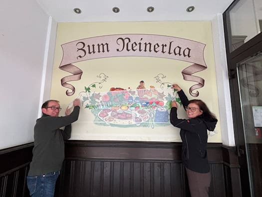 Matthias Singer und Kristin Hängekorb entfernen vorsichtig die Schutzfolie von einem bunten Wandgemälde mit dem Schriftzug „Zum Neinerlaa“.