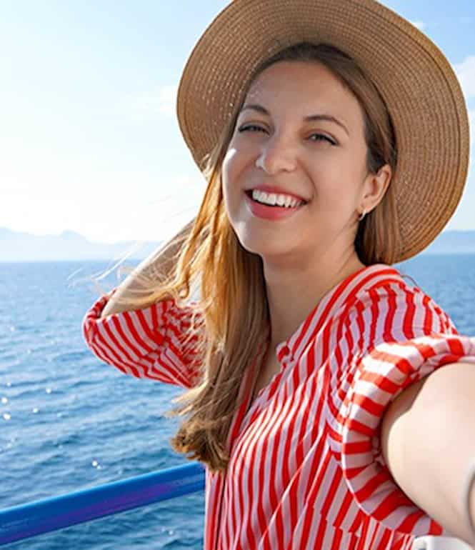 Glücklich lachende junge Frau mit Sonnenhut an Deck eines Schiffs, im Hintergrund das glitzernde Meer und strahlender Sonnenschein.